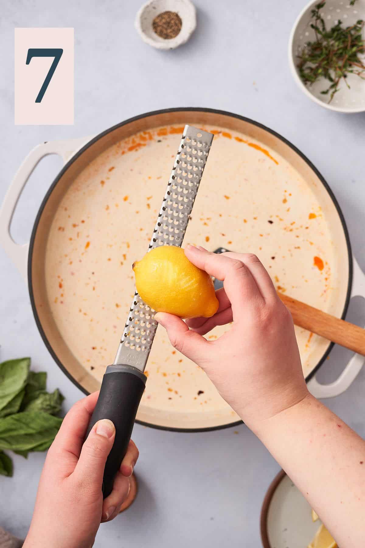 hand zesting a lemon over top of a cream sauce in an enameled cast iron skillet.
