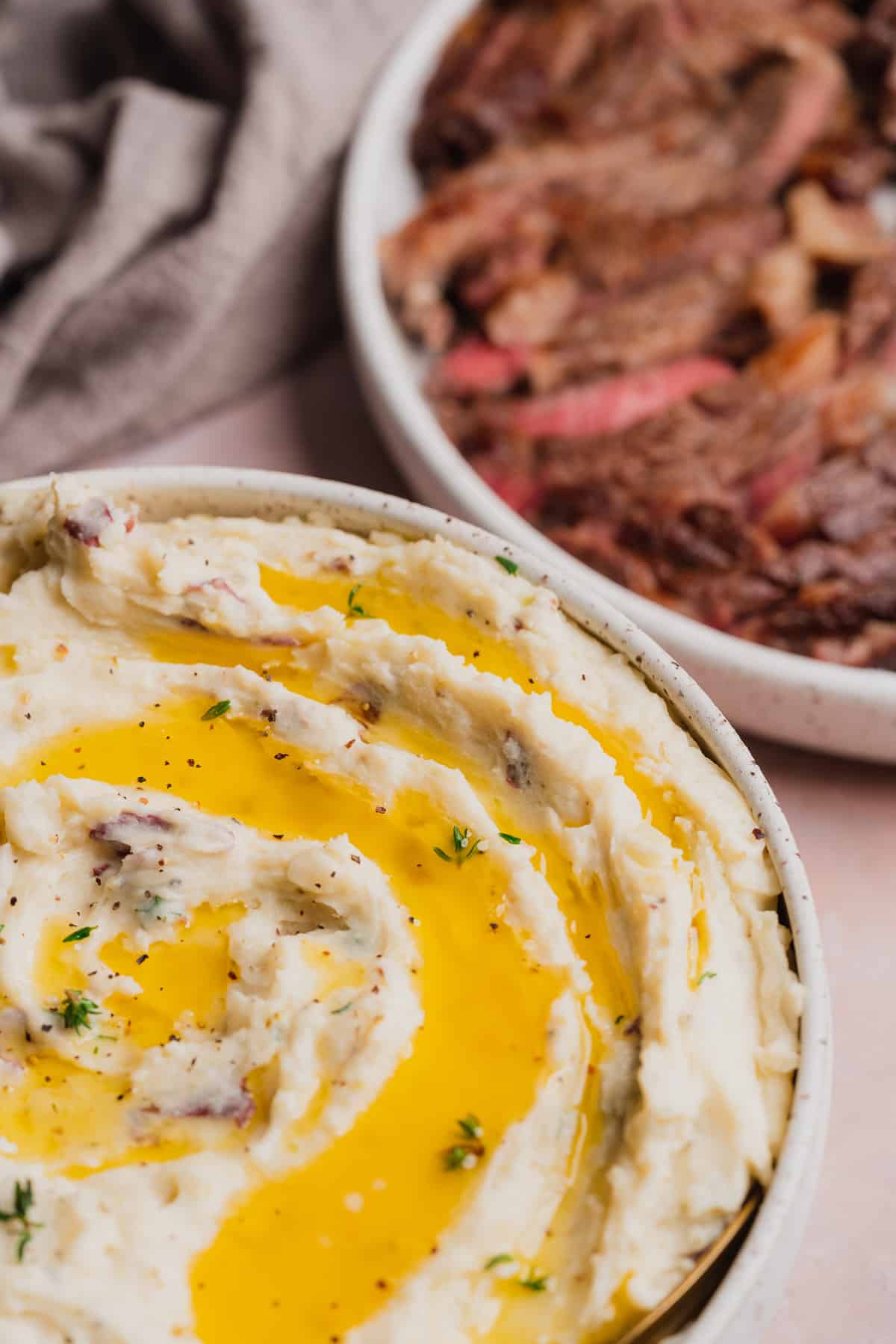 45 degree shot of a bowl of creamy mashed potatoes topped with melted butter, fresh thyme, black pepper, and steak