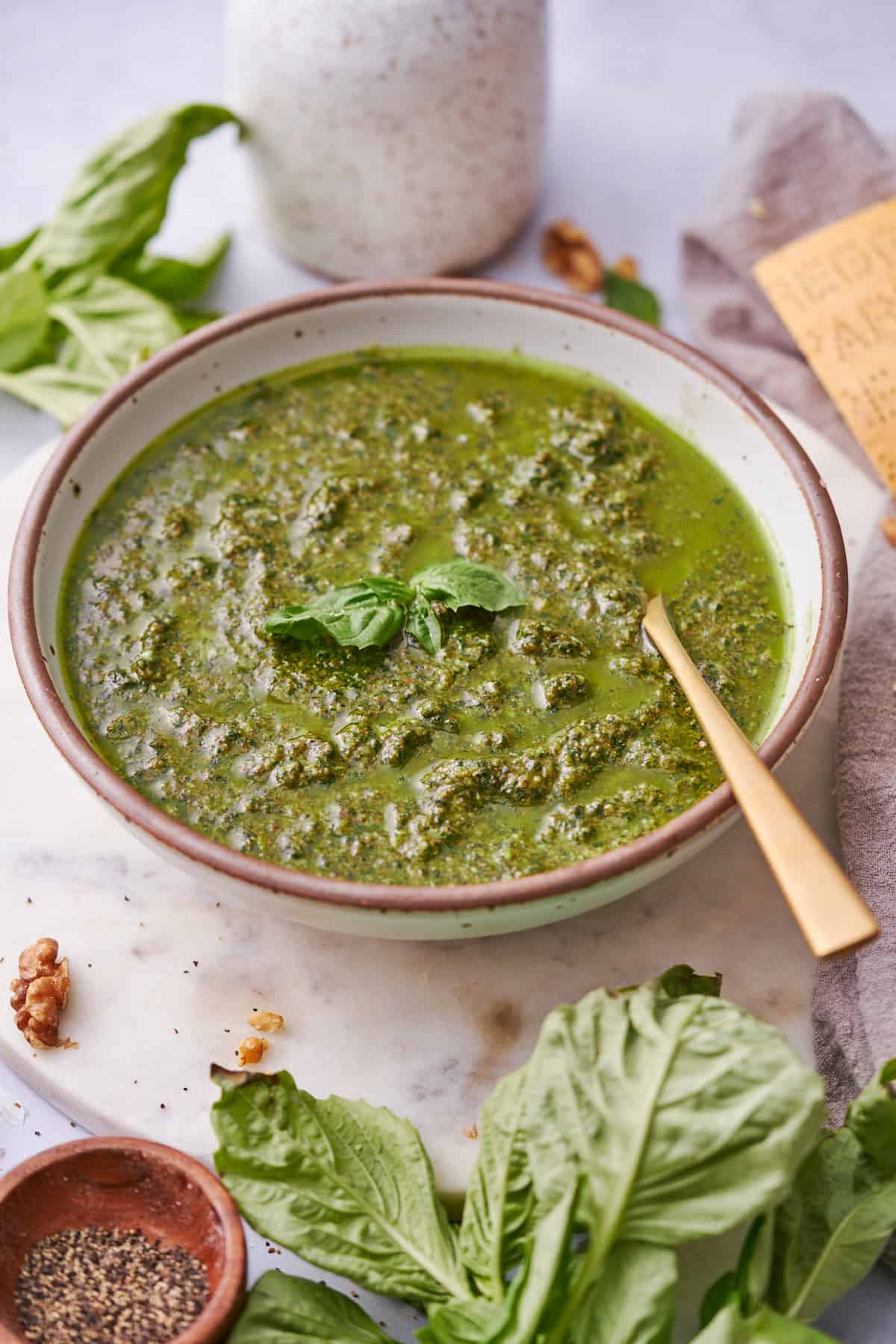 large bowl of freshly made walnut pesto with a gold spoon in the bowl, surrounded by ingredients to make the pesto. 
